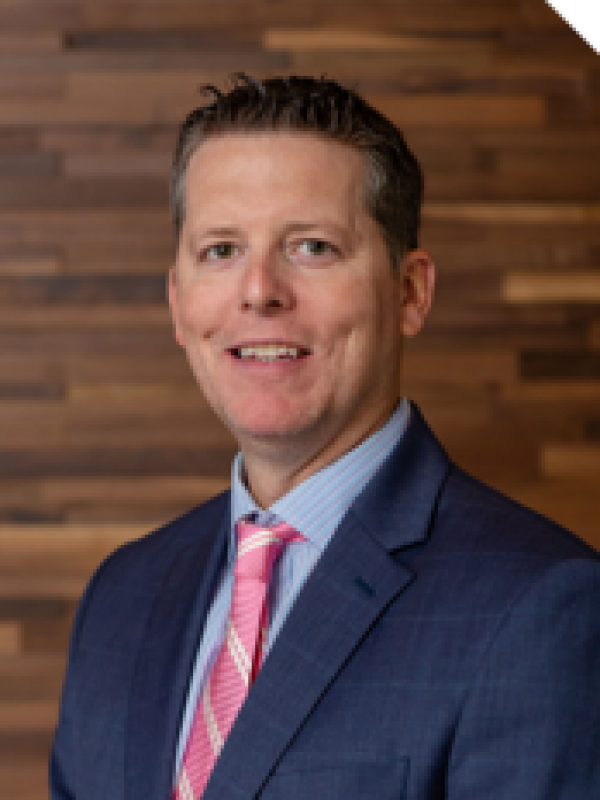 Westrum Corporate John Westrum, in a blue suit and pink tie, smiles as he stands before a wooden wall background.